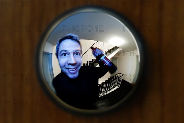 View through door viewer into a staircase of a big tenement, peephole, spyhole, with a man in front of the door grinning and holding a bottle of red wine like a cavalier, admirer, ex-boyfriend