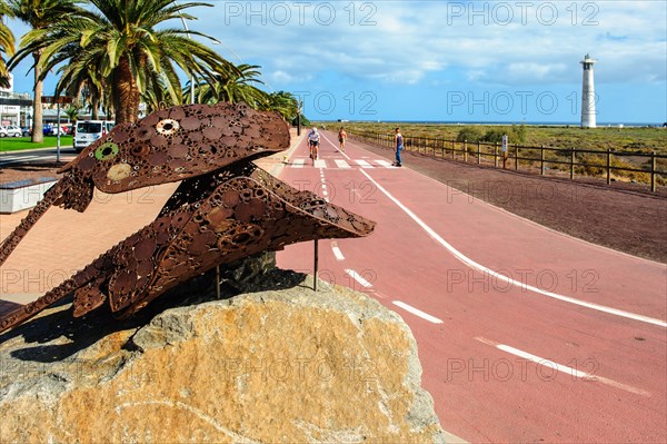 Sculpture made of scrap metal of marine animals two stingrays rays in the left foreground, in the centre of the picture developed cycle path wide cycle road, in the right background lighthouse Faro de Morro Jable, Morro Jable, Jandia peninsula, Fuerteventura, Canary Islands, Canary Islands, Spain, Europe