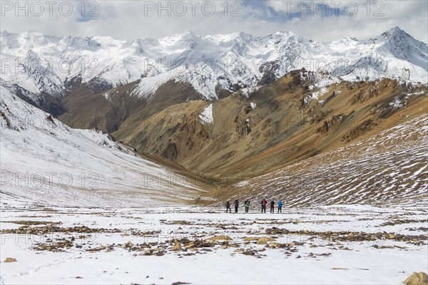 Looking down from Zherin La, a mountain pass in the remote part of the Zanskar Region, towards the Lungnak Valley, with the main ridge of the Great Himalayan Range visible on its other side. A group of trekkers is walking down the slope, towards the distant valley. Photographed in early Autumn, when slopes were covered in fresh snow. Zanskar Range of the Himalayas, dry mountains belonging to the Thetys Himalayas. Kargil District, Union Territory of Ladakh, India, Asia