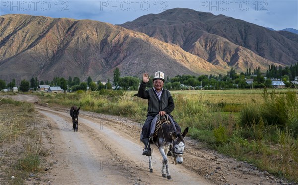 Man in traditional Kyrgyz dress riding on a donkey along a village street and greeting friendly, Kyrgyzstan, Asia
