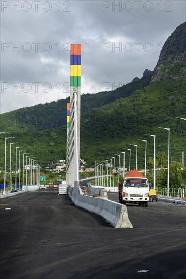 A highway with Mauritian emblem on pillar and a truck driving, surrounded by mountains and an overcast sky.A1-M1 Link Bridge At Grand River North West Valley on the island of Mauritius
