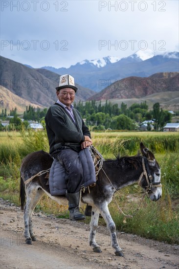 Man in traditional dress riding a donkey in front of a mountain backdrop, Kyrgyzstan, Asia