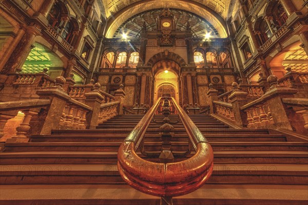 A magnificently illuminated interior staircase in a historic building with a clock in the background, Antwerp-Centraal railway station, Antwerp, Belgium, Europe