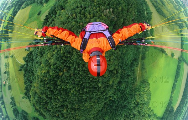 Paraglider pilot with orange clothes and red helmet flying in 500 ft above a forest and meadows, bird's eye view, Brauneck, Lenggries, Bavaria, Germany, Europe