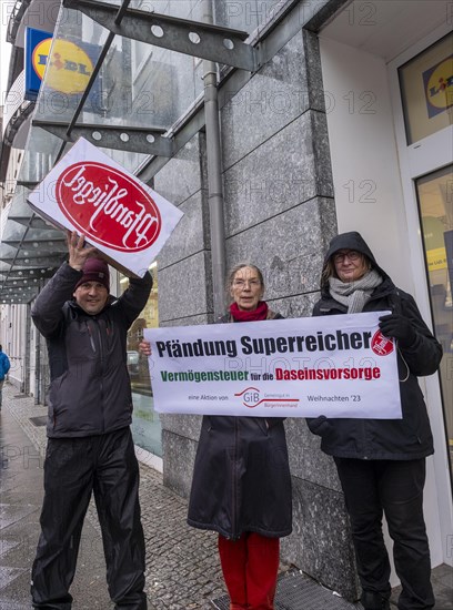 Germany, Berlin, 21.12.2023, action by Gemeingut in BuergerInnenhand (GiB), actions in front of branches of LIDL, Aldi and BMW, here a Lidl branch in Luisenstrasse / Berlin-Mitte, activists press a large deposit seal on a door of the LIDL branch, Carl Wassmuth (left), spokesman for Gemeingut: Super-rich people like LIDL owner Dieter Schwarz only pay tiny tax rates, Europe