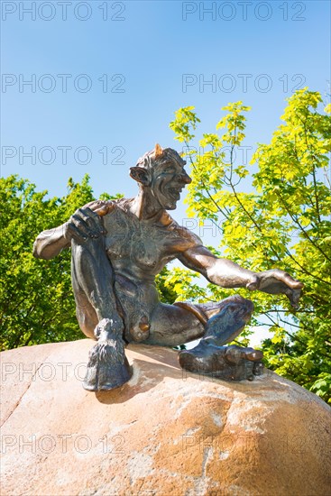 Bronze statue depicting a grinning, laughing naked fantasy figure, placed on a rock, statue, sculpture, sculpture, bronze devil with horns, long ears, hairy shoulders, tail and goat's foot, hoof, on a boulder, part of the group of figures with Homununculus and witch grandmother Wadelinde, Hexentanzplatz, Thale, Eastern Harz, Harz Mountains, Saxony-Anhalt, Germany, Europe