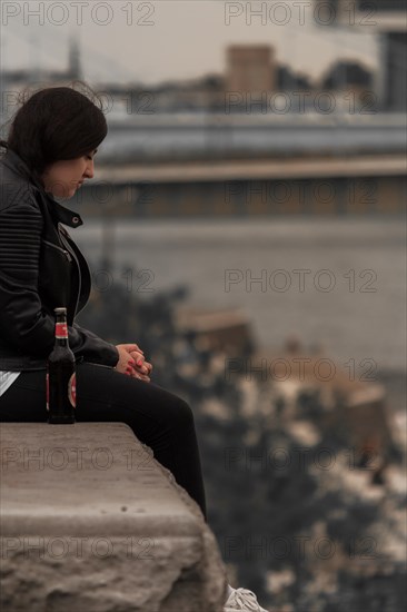 A pensive-looking woman sits on the riverbank with the city skyline in the background, Hohenzollern Bridge, Cologne Deutz, North Rhine-Westphalia, Germany, Europe