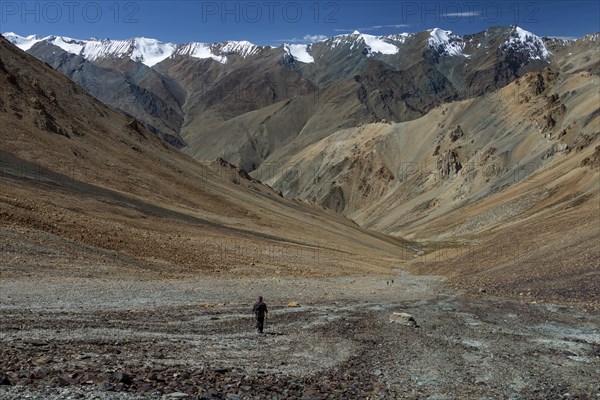 Looking down from Zherin La, a mountain pass in the remote part of the Zanskar Region, towards the Lungnak Valley, with the main ridge of the Great Himalayan Range visible on its other side. A lonely trekker is walking down the slope, and there are a few more below, in a distance. Photographed on a clear, late September day, with clear blue sky above the mountain chain. Zanskar Range of the Himalayas, dry mountains belonging to the Thetys Himalayas. Kargil District, Union Territory of Ladakh, India, Asia