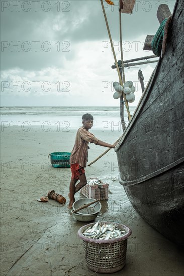 Fisherman by his boat on the beach with the catch of the day during a monsoon shower, Cox's Bazar, Bangladesh, Asia