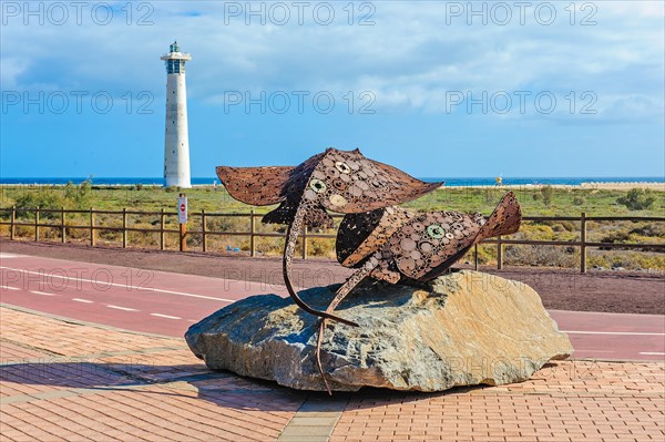 In the foreground sculpture made of scrap metal of marine animals two stingrays rays on stone pedestal, behind it developed cycle path wide cycle road with markings, left in the background lighthouse Faro de Morro Jable, Morro Jable, Jandia peninsula, Fuerteventura, Canary Islands, Canary Islands, Spain, Europe