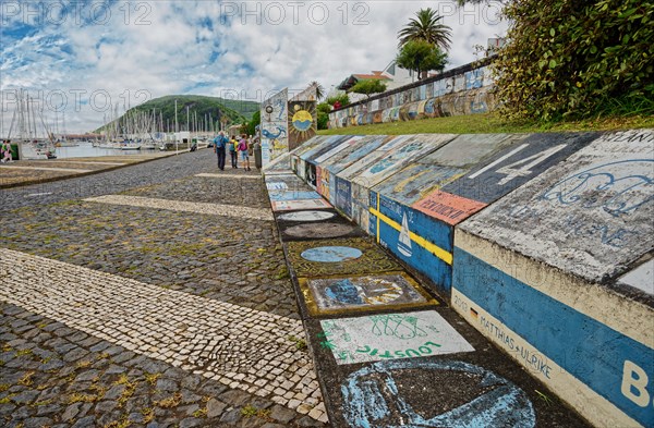 Colourful wall art of Atlantic crossings along a harbour wall with walkers and sailing boats in the background, Horta, Faial Island, Azores, Portugal, Europe