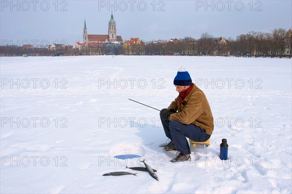 Man with winter cap or hat and warm clothes, sitting on a bucket, ice fishing on Theresienwiese at an ice hole with three mackerels lying beside him in the snow, Theresienwiese is the Oktoberfest Area, Munich, Bavaria, Germany, Europe, it is a joke, because there is no water under the snow, Europe
