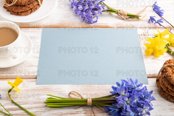 Blue paper sheet with oatmeal cookies, spring snowdrop flowers bluebells, narcissus and cup of coffee on white wooden background. side view, copy space, still life. Breakfast, morning, spring concept