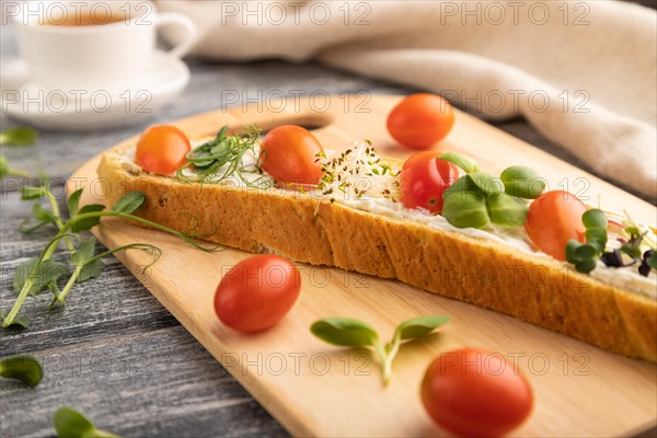 Long white bread sandwich with cream cheese, tomatoes and microgreen on gray wooden background and linen textile. side view, close up, selective focus