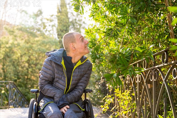 Happy disabled man in wheelchair enjoying while smelling a plant in an urban park during a sunny day of winter