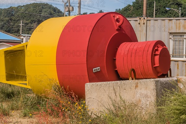 Top view of large yellow and red ocean buoy laying in grass in front of old rusty storage building