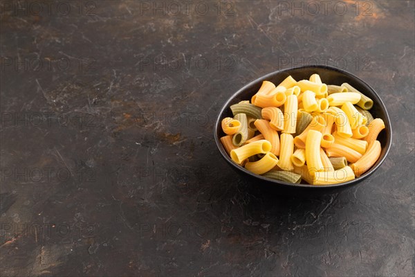 Rigatoni colored raw pasta with tomato, eggs, spices, herbs on black concrete background. Side view, copy space