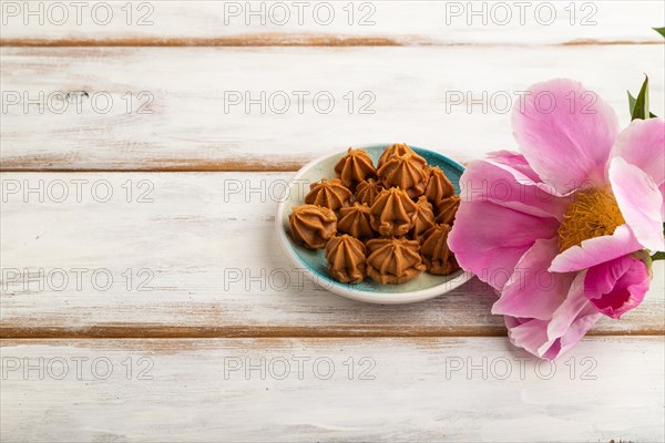 Homemade soft caramel fudge candies on blue plate on white wooden background, peony flower decoration. side view, copy space