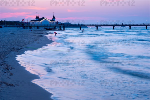 Calm evening mood on the beach with calm waves and two people near a pier with a building at dusk, clear blue-violet sky with pink clouds, Ahlbeck pier in the last evening light, motion blur, wipe effect, long exposure, view from the western beach, Ahlbeck seaside resort, Usedom Island, Mecklenburg-Western Pomerania, Germany, Europe