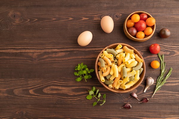 Rigatoni colored raw pasta with tomato, eggs, spices, herbs on brown wooden background. Top view, flat lay, copy space