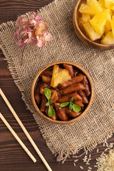 Tteokbokki or Topokki, fried rice cake stick, popular Korean street food with spicy jjajang sauce and pineapple on brown wooden background and linen textile. Top view, flat lay