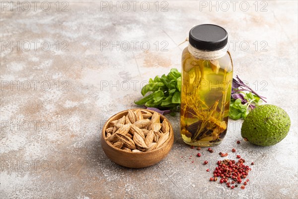 Sunflower oil in a glass jar with various herbs and spices, sesame, rosemary, avocado, basil, almond on a brown concrete background. Side view, copy space