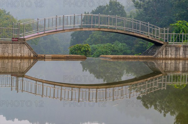 Foot bridge over a small pond with reflection in the water with trees in the background and a dull gray sky in South Korea