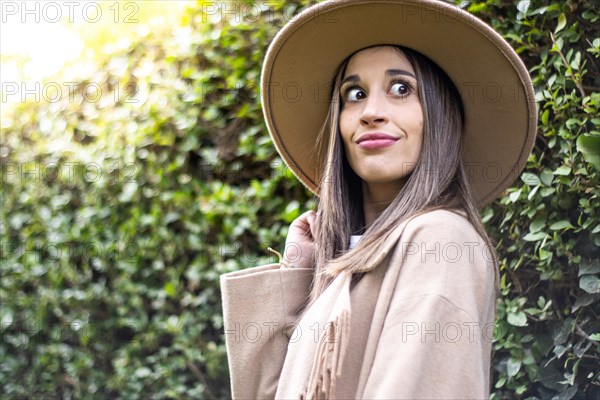 Front view of a young woman in black leather jacket smiling and holding her hat with green leaves in the background outdoors