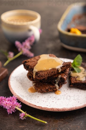 Chocolate brownie with caramel sauce with a cup of coffee on black concrete background. side view, close up, selective focus