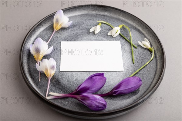 White paper invitation card, mockup with crocus and galanthus flowers on ceramic plate and gray background. Blank, side view, still life, copy space, wedding invitation