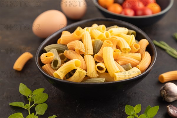 Rigatoni colored raw pasta with tomato, eggs, spices, herbs on black concrete background. Side view, close up, selective focus