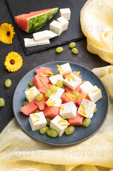 Vegetarian salad with watermelon, feta cheese, and grapes on blue ceramic plate on black concrete background and yellow linen textile. Side view, close up