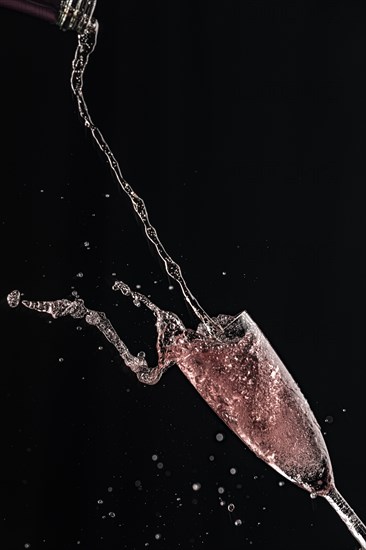 Rose champagne pours from a bottle into a champagne glass, drops and splashes fly, black background