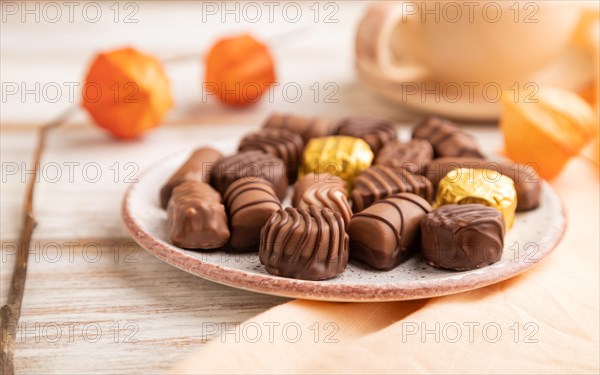 Chocolate candies with cup of coffee and physalis flowers on a white wooden background and orange textile. side view, close up, selective focus