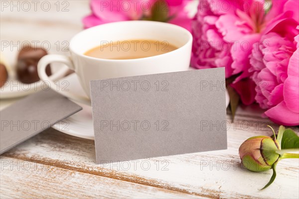 Gray business card with pink peony flowers and cup of coffee on white wooden background. side view, copy space, still life. Breakfast, morning, spring concept