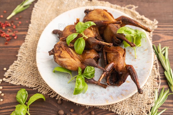 Smoked quails with herbs and spices on a ceramic plate with linen textile on a brown wooden background. Side view, close up