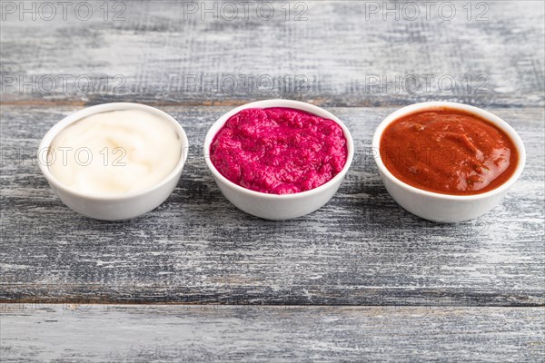 Ceramic sauce bowls with horseradish, tomato sauce and mayonnaise on a gray wooden background. Side view, close up, copy space