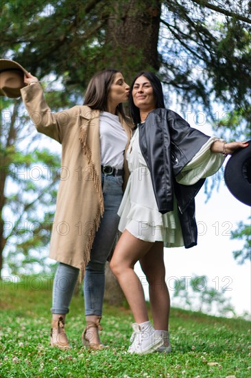 Lifestyle of two girls with hats kissing each other on the cheek in the grass. Two happy best friends in hats kissing cheeks outdoors. Girls friendship concept