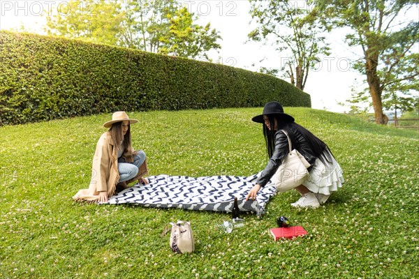 Side view of a two stylish friends spreading a blanket on the lawn of a park to spend the day together there