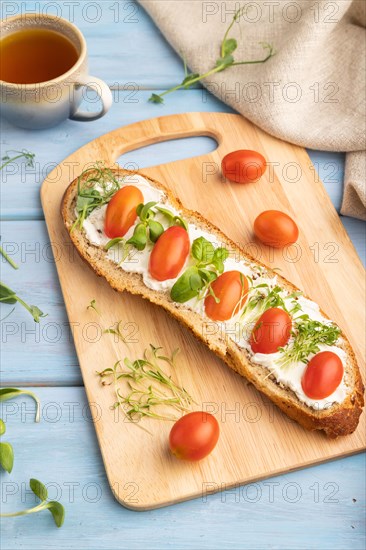 Long white bread sandwich with cream cheese, tomatoes and microgreen on blue wooden background and linen textile. side view, close up