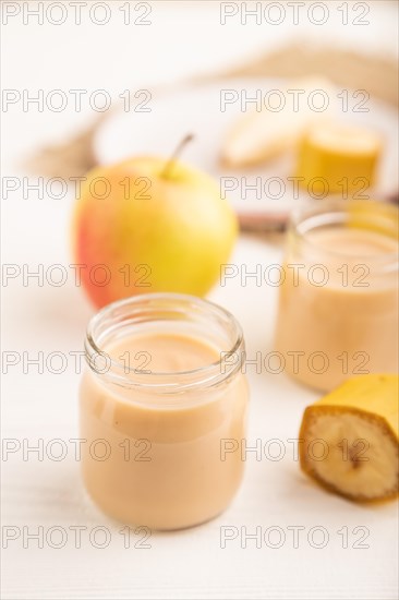 Baby puree with fruits mix, apple, banana infant formula in glass jar on white wooden background. Side view, close up, selective focus, artificial feeding concept