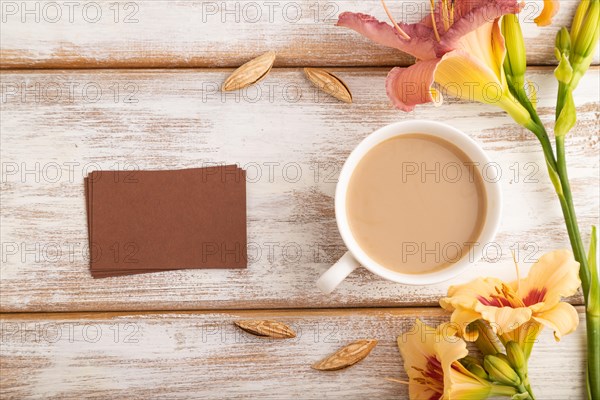 Brown paper business card mockup with orange day-lily flower and cup of coffee on white wooden background. Blank, top view, flat lay, copy space, still life. spring concept