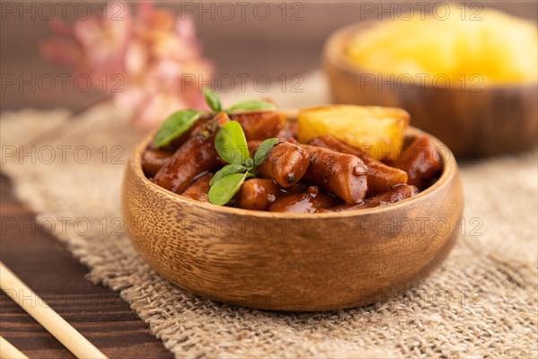 Tteokbokki or Topokki, fried rice cake stick, popular Korean street food with spicy jjajang sauce and pineapple on brown wooden background and linen textile. Side view, selective focus