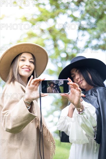 Vertical front view of two stylish friends both grabbing a cell phones to take a selfie