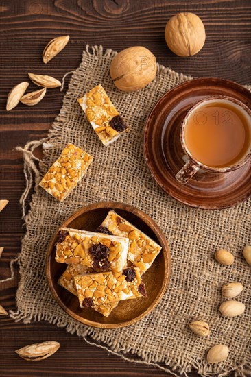 Traditional candy nougat with nuts and sesame with cup of green tea on brown wooden background and linen textile. top view, flat lay, close up
