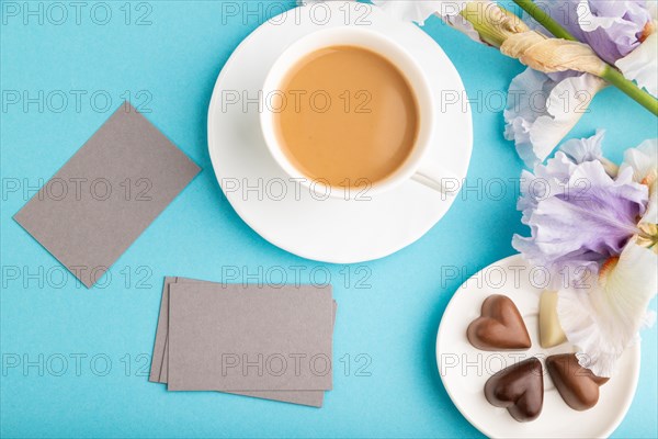 Gray business card with cup of cioffee, chocolate candies and iris flowers on blue pastel background. top view, flat lay, copy space, still life. Breakfast, morning, spring concept