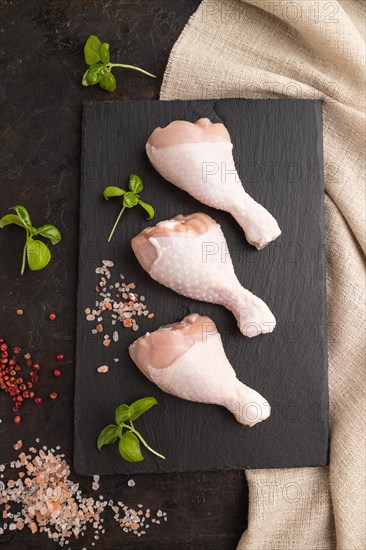 Raw chicken legs with herbs and spices on a black slate cutting board on a black concrete background and linen textile. Top view, flat lay, close up