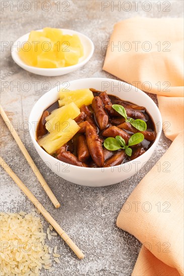 Tteokbokki or Topokki, fried rice cake stick, popular Korean street food with spicy jjajang sauce and pineapple on gray concrete background and orange textile. Side view, close up