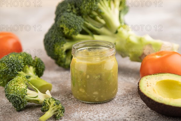 Baby puree with vegetable mix, broccoli, tomatoes, cucumber, avocado infant formula in glass jar on brown concrete background. Side view, close up, selective focus, artificial feeding concept