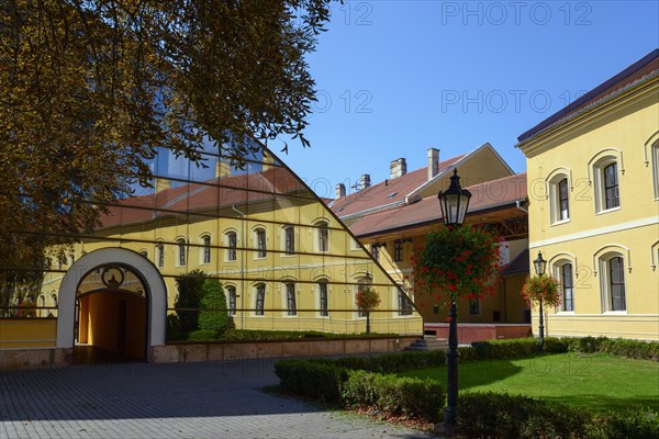 A yellow building reflected in a large glass facade, flanked by trees and a paved path, Officers' Palace, Komarno, Komarom, Komorn, Nitriansky kraj, Slovakia, Europe